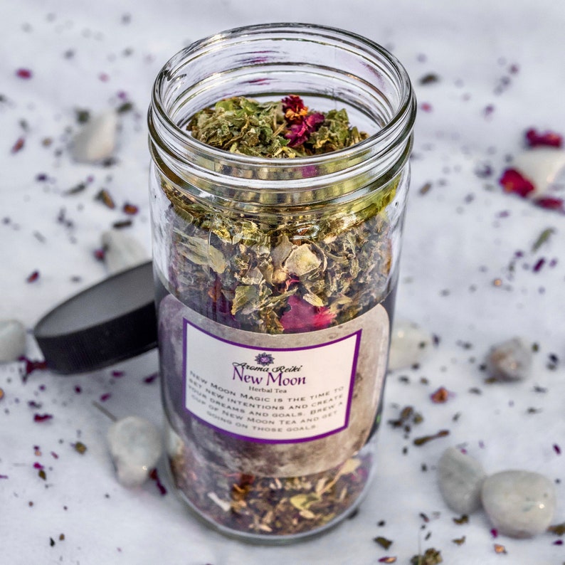 May include: A glass jar filled with a blend of dried herbs and flowers. The label reads "Aroma Reiki Herbal Tea New Moon" and includes the text "New Moon Magic is the time to set new intentions and create new dreams and goals. Brew a cup of New Moon Tea and get going on those goals."