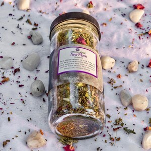 May include: A glass jar filled with a blend of dried herbs and flowers, labeled "New Moon Herbal Tea". The label reads "New Moon Magic is the time to set new intentions and create your dreams and goals. Brew a cup of New Moon Tea and get going on those goals."