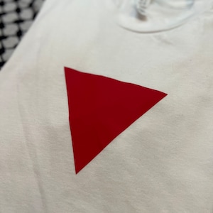 May include: A white t-shirt featuring a large, solid red triangle in the center. The shirt is made of a soft material and is folded. The background is a black and white patterned fabric.