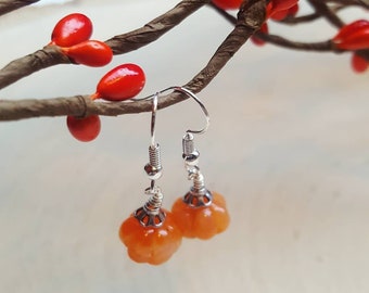 May include: A pair of dangle earrings featuring small, orange, pumpkin-shaped beads. The earrings have silver-colored hooks and decorative metal caps. The background includes a branch with red berries, creating a seasonal or autumnal theme.