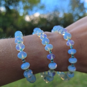 May include: Three beaded bracelets with opalescent, light blue beads and smaller teal beads. The bracelets are worn on a wrist, with a blurred background of green foliage and a blue sky.