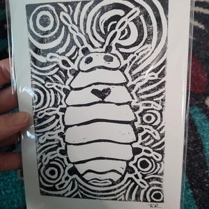 May include: Black and white linocut print of a bug with a heart on its chest. The bug is surrounded by concentric circles.