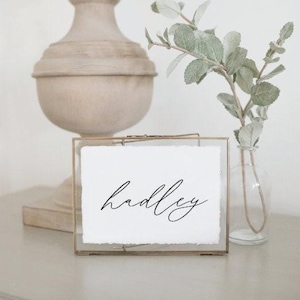 May include: A clear glass frame holds a white paper with the name "hadley" written in black cursive script. A decorative lamp and a vase with greenery are in the background. The frame is on a light gray surface.