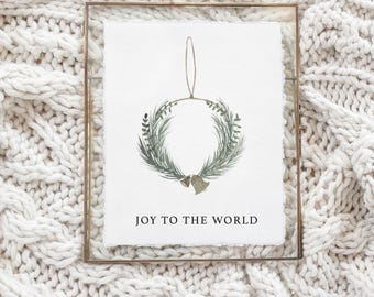 May include: A framed watercolor print featuring a Christmas wreath with green foliage and two small bells. The text "JOY TO THE WORLD" is printed below the wreath. The frame is a thin, gold-colored metal.