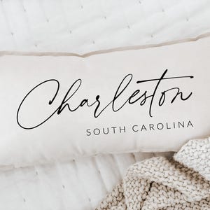 May include: Cream-colored rectangular pillow with the word "Charleston" in elegant black script. Below, the text "SOUTH CAROLINA" is printed in a smaller, sans-serif font. The pillow is resting on a white surface with a knitted blanket.