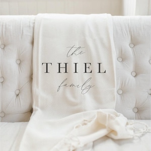 K&ouml;nnte beinhalten: Cremefarbene Decke, drapiert &uuml;ber einem wei&szlig;en, getufteten Sofa. Die Decke tr&auml;gt den Text "the THIEL family" in einer stilvollen Schriftart. Die Decke hat ein strukturiertes Gewebe und einen Fransenrand, der einen Hauch von Eleganz verleiht.