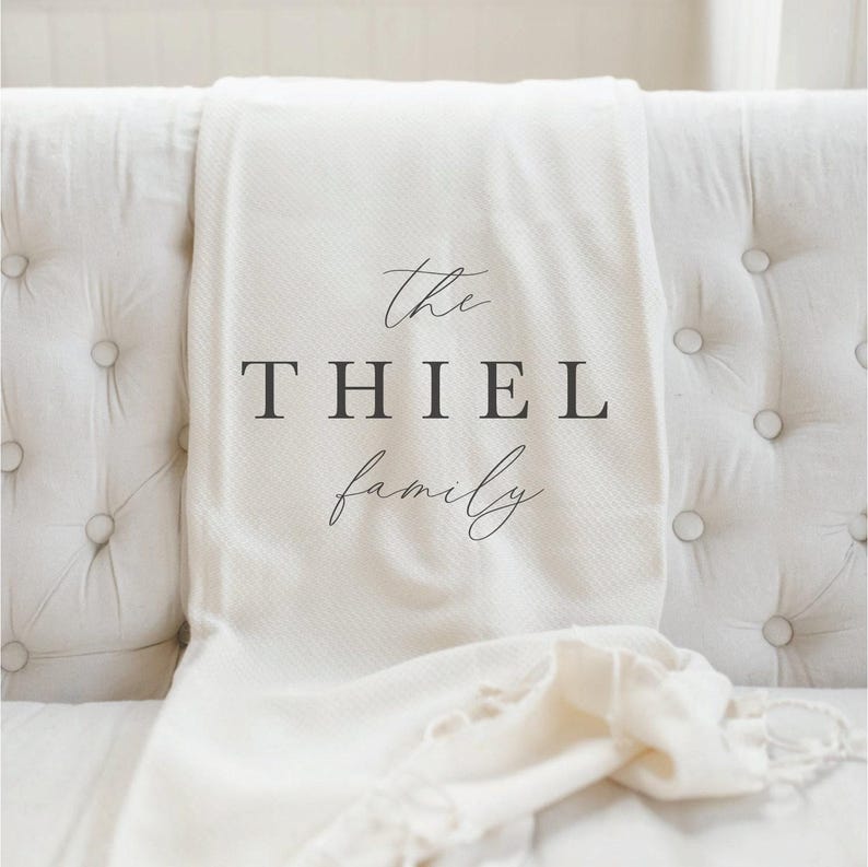 K&ouml;nnte beinhalten: Cremefarbene Decke, drapiert &uuml;ber einem wei&szlig;en, getufteten Sofa. Die Decke tr&auml;gt den Text "the THIEL family" in einer stilvollen Schriftart. Die Decke hat ein strukturiertes Gewebe und einen Fransenrand, der einen Hauch von Eleganz verleiht.