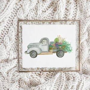 May include: Watercolor painting of a vintage gray pickup truck filled with potted flowers. The truck bed is overflowing with purple, green, and yellow blooms. The artwork is framed in a thin, gold-colored metal frame.