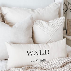 May include: A collection of ivory-colored decorative pillows with varying textures and designs. One rectangular pillow displays the word "WALD" above "est. 2018" in black script. The pillows rest on a bed with a knitted throw.