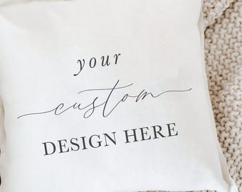 Könnte beinhalten: Cremefarbenes quadratisches Kissen mit den Wörtern "your custom DESIGN HERE" in Schwarz aufgedruckt. Es liegt auf einer beigefarbenen Strickdecke. Das Kissen hat ein schlichtes, elegantes Design.