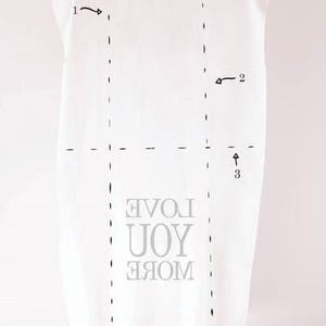 K&ouml;nnte beinhalten: Rechteckige wei&szlig;e Decke mit dem Text "LOVE YOU MORE" in Grau. Die Decke hat einen Fransen am unteren Rand. Die Decke wird von einer Person gehalten.