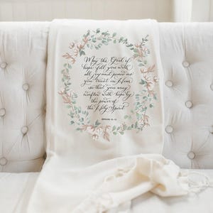 May include: Cream-colored throw blanket draped over a white tufted sofa. The blanket features a floral wreath design with text that reads "May the God of hope fill you with all joy and peace as you trust in Him..." Romans 15:13.