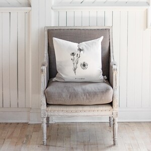 May include: A white throw pillow with a black botanical illustration of a flower and the text "No. 3 sunflower" rests on a gray upholstered chair. The chair has a distressed, light-colored wooden frame and is set against a white paneled wall.