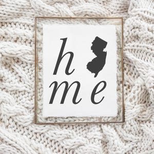 May include: A framed print with the word "home" and a silhouette of New Jersey in black. The print is set against a white background and framed in a gold-colored metal frame. The background is a white knitted blanket.