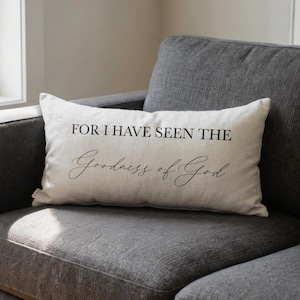May include: Cream-colored rectangular pillow with the text "FOR I HAVE SEEN THE Goodness of God" in black script. The pillow is resting on a gray sofa. The pillow has a simple, elegant design.