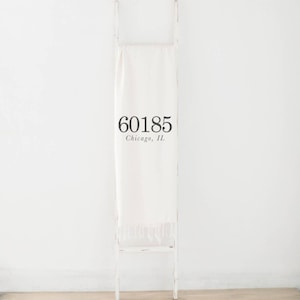 May include: A white throw blanket with the text "60185 Chicago, IL" draped over a white decorative ladder. The blanket has a fringed edge and is displayed against a white wall.