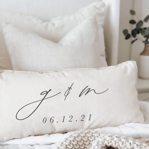 May include: Cream-colored rectangular pillow with black script initials and the date 06.12.21. The pillow is on a white bed with other pillows and a knitted blanket. The initials are in a cursive font.