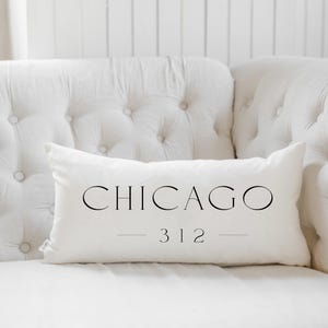 May include: A white rectangular pillow with the word "CHICAGO" in black, elegant font. Below the city name is the number "312". The pillow is resting on a white tufted sofa.