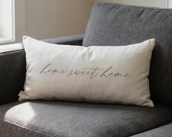 Könnte beinhalten: Ein rechteckiges, cremefarbenes Kissen mit dem Schriftzug "home sweet home" in Schreibschrift. Das Kissen liegt auf einem grauen Sofa. Der Stoff hat ein weiches, strukturiertes Aussehen.