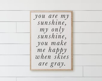 May include: A rectangular wall sign with a light-colored background and a thin wooden frame. The sign features black cursive text that reads: you are my sunshine, my only sunshine, you make me happy when skies are gray. The sign is oriented vertically.