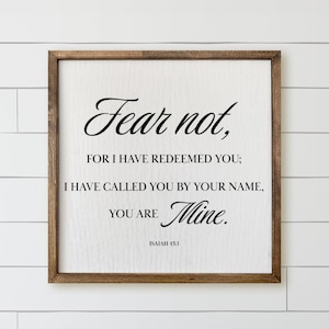 May include: A square wooden sign with a dark brown frame and a white background. The text in black reads: Fear not, FOR I HAVE REDEEMED YOU; I HAVE CALLED YOU BY YOUR NAME, YOU ARE Mine. ISAIAH 43:1. The sign features a visible wood grain texture.