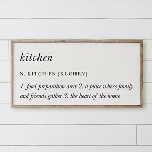 May include: A rectangular wooden framed sign with the word "kitchen" in a black, elegant font. Below the word is a definition, including the pronunciation and three descriptive points about the kitchen. The sign is mounted on a white, horizontal plank wall.