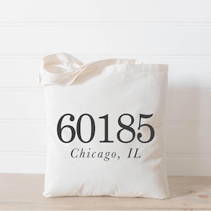 May include: Cream-colored tote bag with black text. The text reads "60185 Chicago, IL". The bag has a simple design and is made of a soft, foldable material. The bag has a handle for carrying.