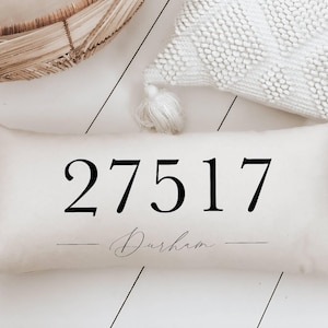 May include: Cream-colored rectangular pillow with the numbers "27517" printed in black. Below the numbers, the word "Durham" is written in a cursive font. The pillow is a decorative home accent.
