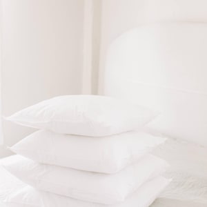 May include: A stack of four white square pillows on a bed. The pillows are fluffy and appear to be made of a soft material. The background is a light, neutral color, creating a clean and minimalist aesthetic.