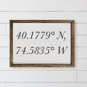May include: A framed rectangular sign with a white background and dark brown wooden frame. The sign displays the coordinates 40.1779° N, 74.5835° W in a black, elegant font. The sign is mounted on a white, horizontal shiplap wall.
