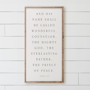May include: A rectangular wall sign with a light wood frame and a white background featuring black text that reads: AND HIS NAME SHALL BE CALLED WONDERFUL, COUNSELOR, THE MIGHTY GOD, THE EVERLASTING FATHER, THE PRINCE OF PEACE. ISAIAH 9:6.