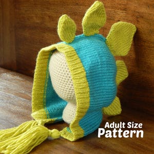 May include: A teal blue and yellow knitted hat with a dinosaur design. The hat has a long yellow tassel and is designed for adults. The text "Adult Size Pattern" is visible in the image.