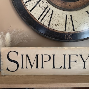 May include: A wooden sign with the word "SIMPLIFY" in black lettering. The sign is rectangular and has a light-colored wood grain. A large clock and a vase with dried flowers are in the background.