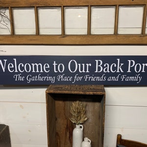 May include: A dark blue wooden sign with white lettering that reads "Welcome to Our Back Porch The Gathering Place for Friends and Family".