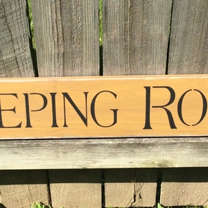 May include: A wooden sign with the words "KEEPING ROOM" painted in black letters on a light brown background.