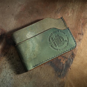 Op de afbeelding: Handgemaakte, verweerde groene leren kaarthouder met een bi-fold ontwerp. De voorkant heeft een reliëflogo met de tekst "Harbinger Leather Design, Messenger of Secrets and Dreams". De randen zijn gestikt met een contrasterende draad.