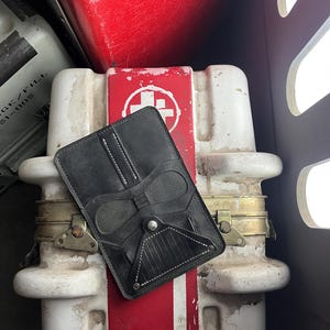 Könnte beinhalten: Ein schwarzer Taschenorganizer aus Leder mit vertikalem Steppmuster und einer unteren Klappe, die mit einem silbernen Metalldruckknopf gesichert ist. Der Organizer hat eine rechteckige Form mit abgerundeten Ecken und weißen Kontrastnähten.