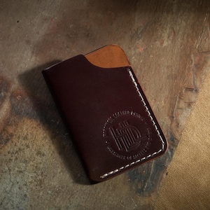 Op de afbeelding: Een donkerbruine leren kaarthouder met een lichtere bruine zak. De kaarthouder heeft witte stiksels rond de randen en een cirkelvormig reliëflogo. Het product is waarschijnlijk handgemaakt.