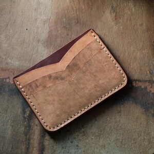 May include: A handmade leather card wallet in shades of brown and burgundy. The wallet features a V-shaped design on the front and is stitched with white thread. The wallet is rectangular and appears to be new.