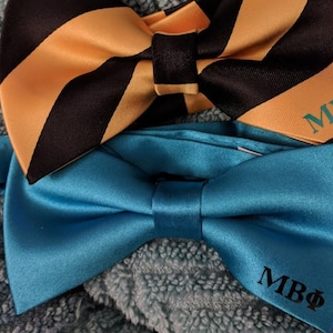 May include: Two bow ties, one black and gold striped and one solid teal blue. Both bow ties have the letters "MBΦ" embroidered on them.