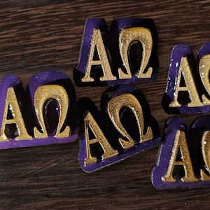 May include: Five purple and gold decorative tiles with the letters "AΩ" on each. The tiles are made of a smooth, glossy material and have a slightly raised design.