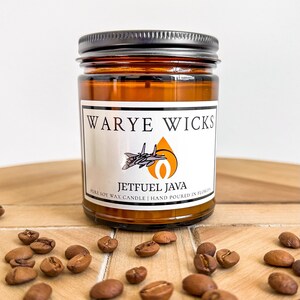Op de afbeelding: Een bruine glazen kaars pot met een zwarte deksel. Het label vermeldt "Warye Wicks Jetfuel Java Pure Soy Wax Candle | Hand Poured in Florida". Een klein vliegtuig is afgebeeld op het label. Koffiebonen zijn verspreid over een houten oppervlak.