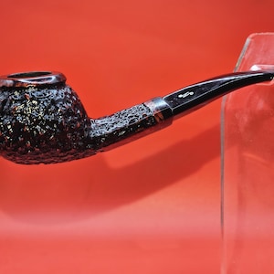 May include: A tobacco pipe with a dark, textured bowl and a sleek, black stem. The pipe is held by a clear acrylic stand against a red background. The stem has a silver band and a small, stylized design.