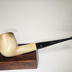 May include: A tobacco pipe featuring a light beige bowl with the text D.E. COLOR stamped on the side. The pipe has a long, smooth black stem with a small yellow dot accent near the junction where the stem meets the bowl, creating a contrast.