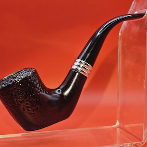 May include: A dark brown tobacco pipe with a textured bowl and a curved, glossy black stem. The pipe features silver bands near the stem's connection to the bowl. The pipe is displayed on a clear stand against a red background.