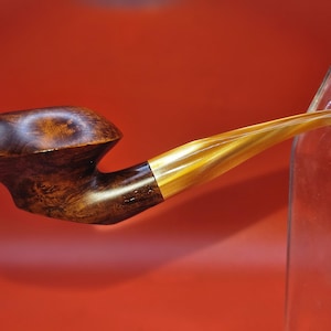 Pode incluir: Um cachimbo de tabaco artesanal com uma piteira curva castanha escura e um bocal âmbar claro. O cachimbo tem uma faixa de latão que conecta a piteira e o bocal. O cachimbo é exibido em um fundo vermelho.
