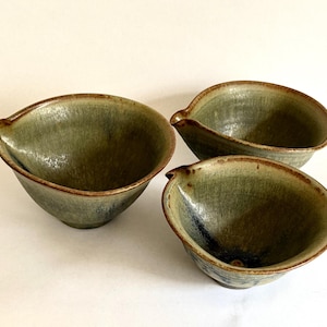 May include: Three ceramic bowls with a glossy, mottled olive green glaze and a distinct pouring spout on one side. Each bowl features a rounded shape with a slightly uneven rim and a dark, textured finish that transitions into lighter green tones.