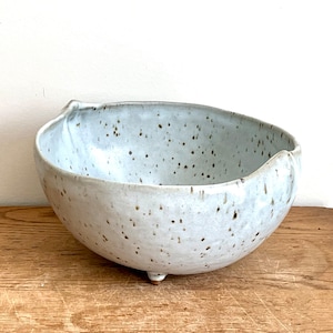 May include: A white ceramic bowl with a speckled glaze and a slightly irregular rim. The bowl has three small feet and is sitting on a wooden surface.