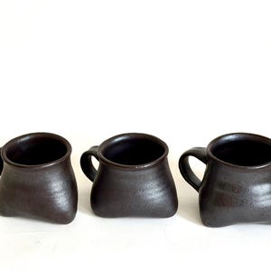 May include: Three dark brown ceramic mugs with a rounded, wide-bottomed shape and a smooth, slightly glossy finish. Each mug features a small, curved handle on the side and a wide, open top, showcasing a consistent dark, earthy tone throughout.