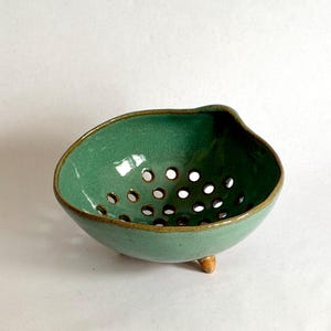 May include: A handmade ceramic colander bowl with a glossy sage green glaze and a dark brown rim. The bowl features a series of circular drainage holes at the bottom and is supported by three small, rounded, light brown clay feet at the base.
